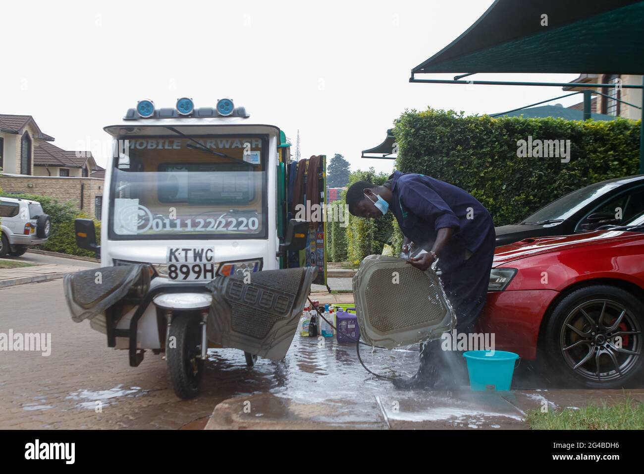 Un membre du personnel de Wash Me Please Mobile Carwash utilise une machine  de lavage de voiture installée dans un Tuk électrique pour nettoyer les  tapis de pied à la maison d'un
