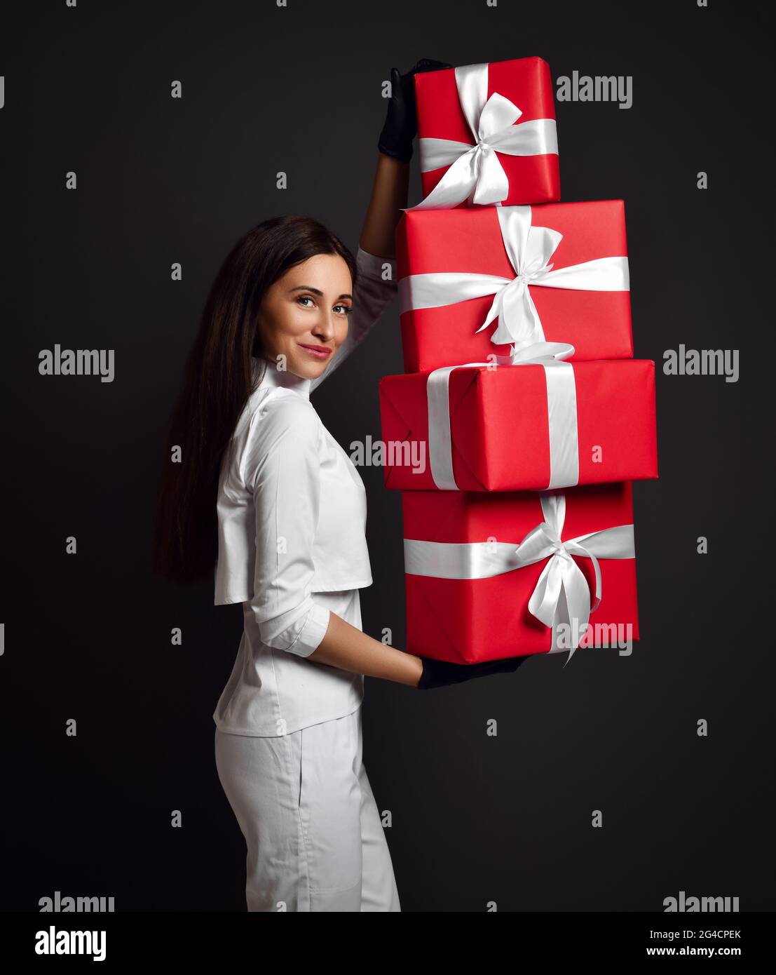 Attrayante jeune femme positive médecin dentiste en uniforme blanc et gants en latex debout et tenant tas de boîtes-cadeaux de vacances Banque D'Images