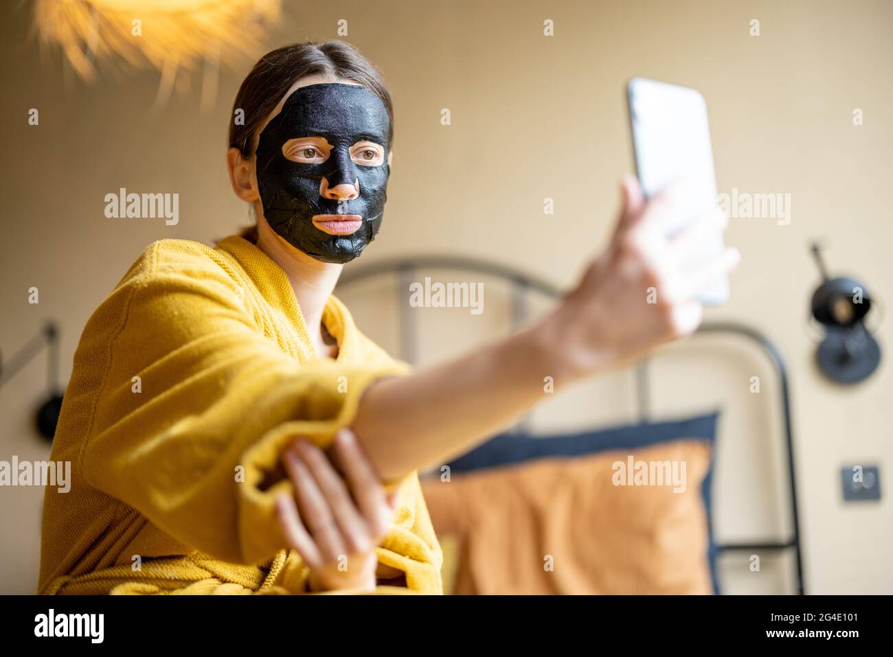 Femme en peignoir avec masque de nettoyage noir et téléphone dans la chambre Banque D'Images