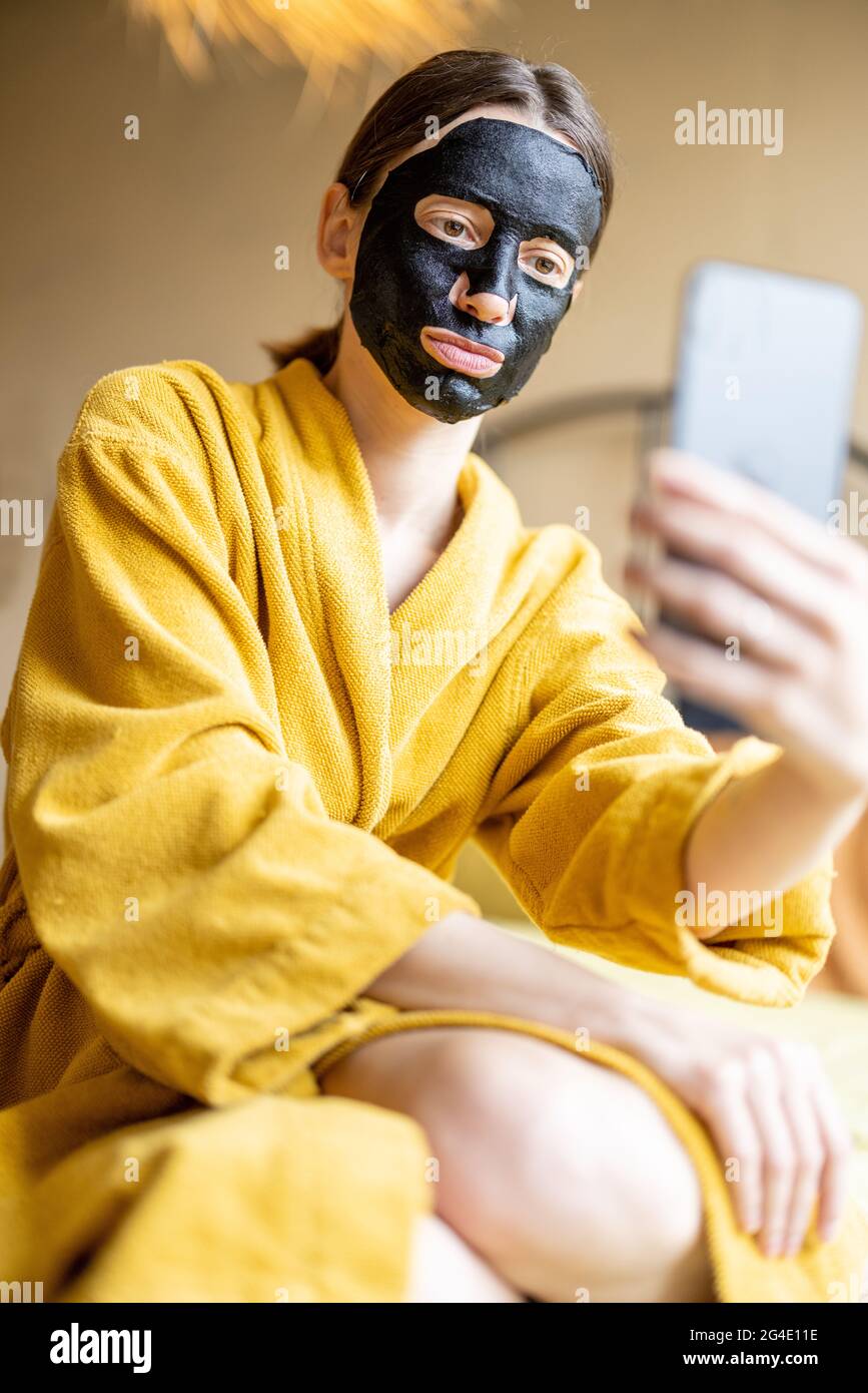 Femme en peignoir avec masque de nettoyage noir et téléphone dans la chambre Banque D'Images