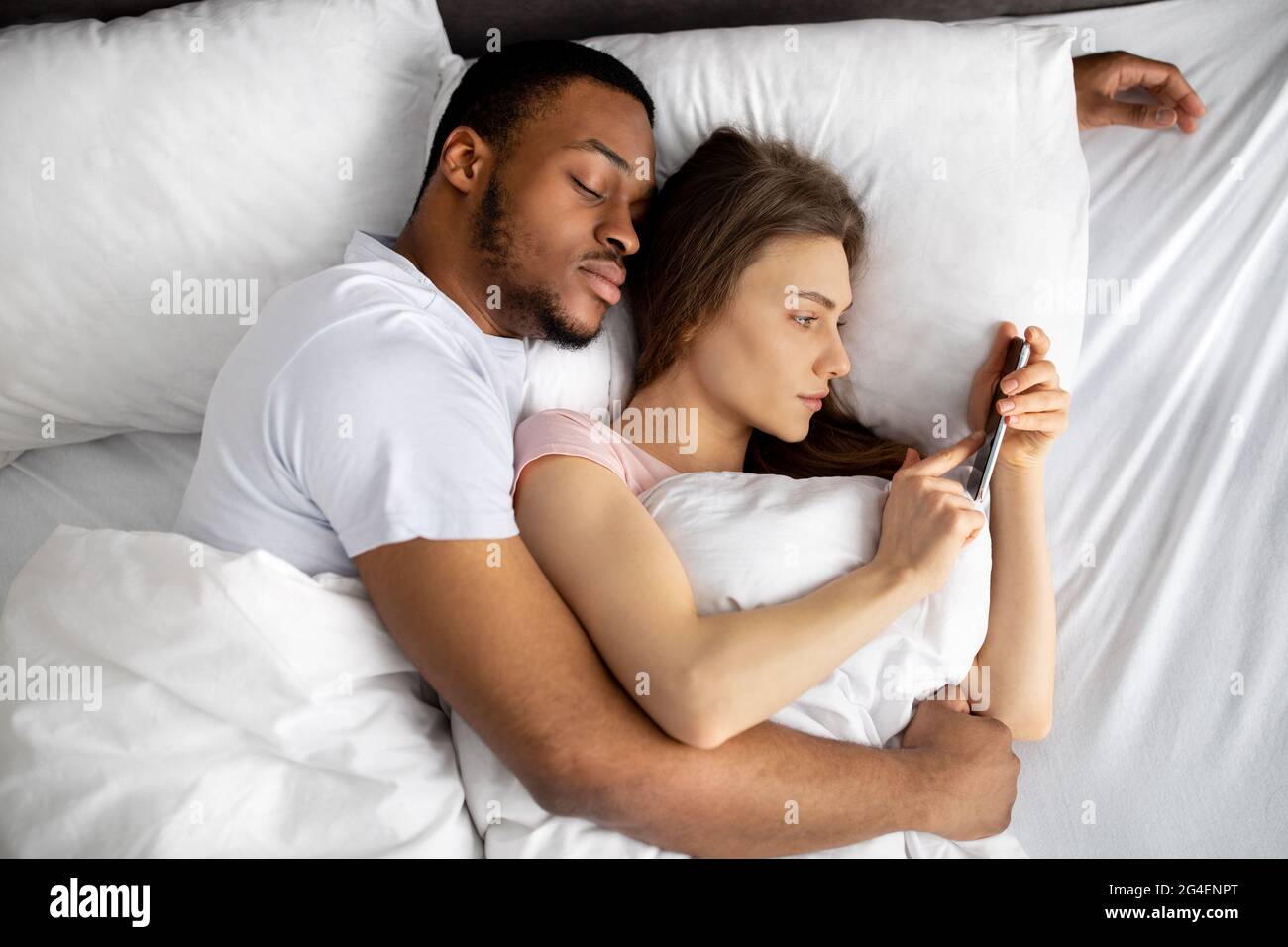 Jeune femme couché sur le lit dans les bras de son mari noir et utilisant le téléphone portable pour envoyer des SMS à son amant, vue du dessus Banque D'Images