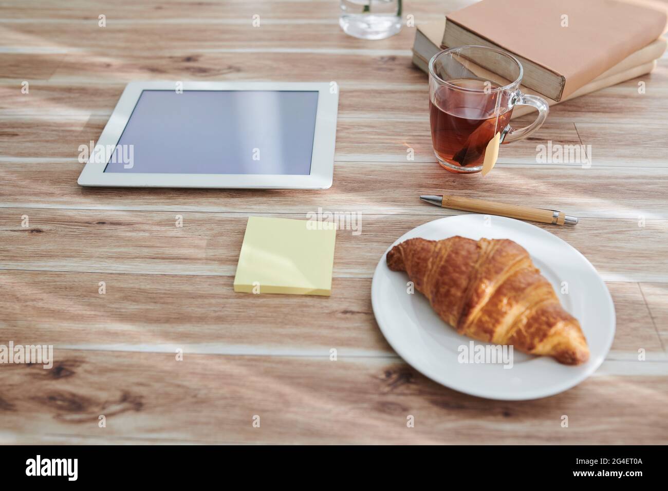 Tasse avec sac de thé et croissant sur l'assiette pour le petit déjeuner à côté de l'ordinateur tablette et pile de livres sur la table de la personne Banque D'Images