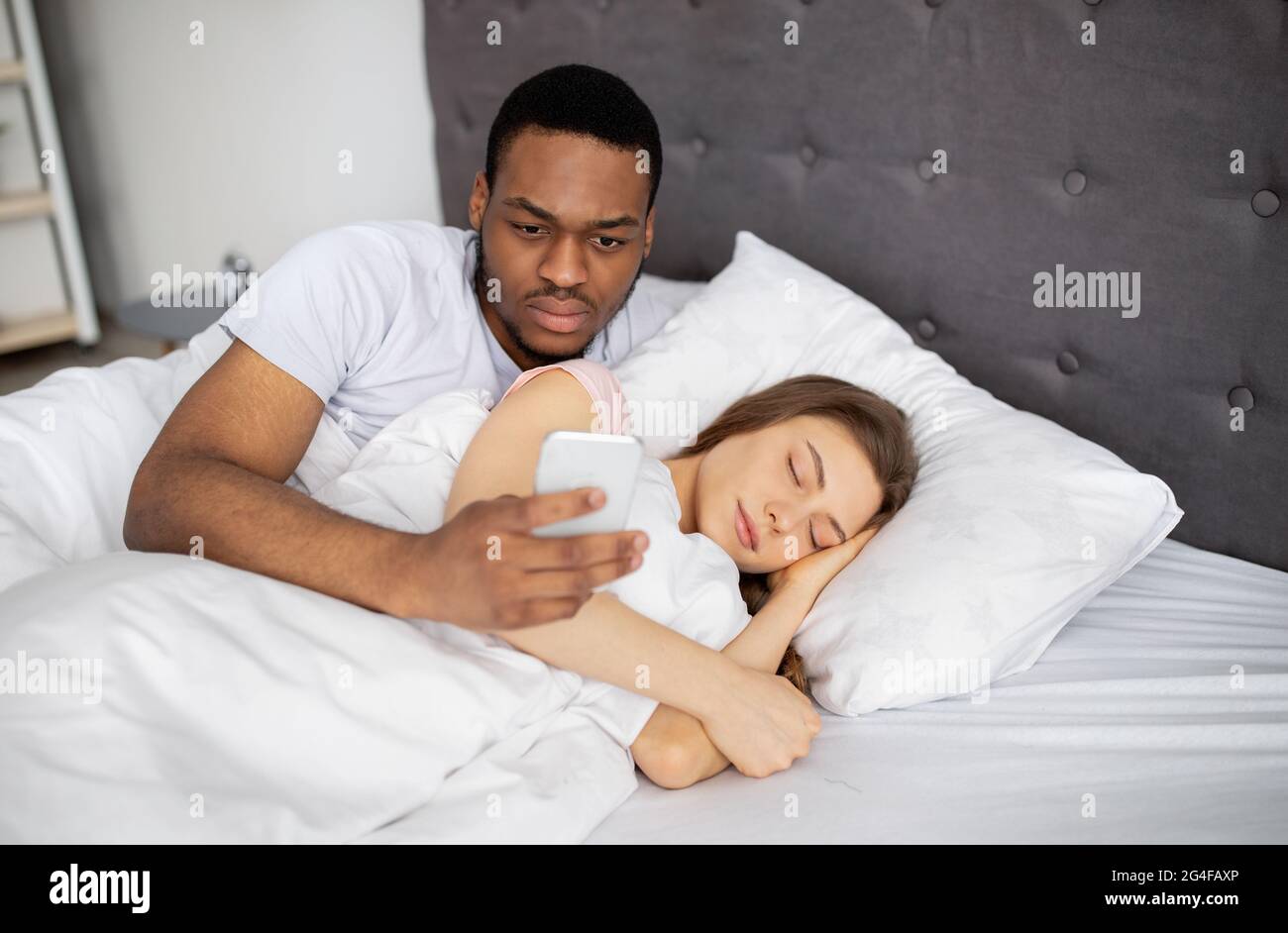 Jaloux mari noir vérifiant le smartphone de sa femme, lisant la correspondance pendant qu'elle est endormi au lit à la maison Banque D'Images
