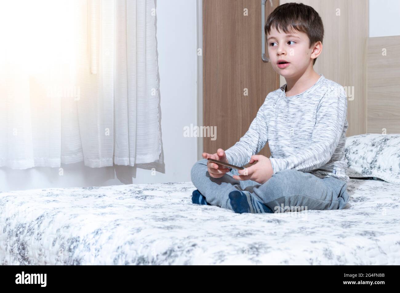 Un petit garçon de 6 ans utilise et joue sur son smartphone. Mignon petit garçon jouant à des jeux sur smartphone dans le lit. Garçon joue dans un jeu vidéo sur son smartphone. Banque D'Images