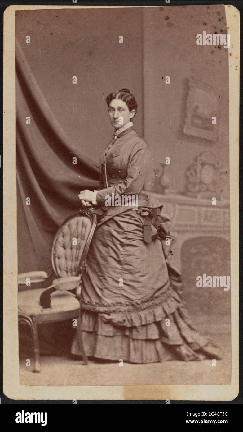 Carte-de-visite d'Anna M. Stanton en portrait complet. Stanton est debout avec son corps tourné vers le côté et son côté gauche avant. Sa tête est tournée vers la caméra. Elle porte ses cheveux séparés au centre et rassemblés dans un updo derrière sa tête. Elle a sur un bodyce de couleur foncée et une jupe d'agitation. Une chatelaine ou une chaîne de montre est visible pendre de sa poitrine en dessous de son bras gauche. Elle est debout devant un décor intérieur peint avec un salon et des draperies. Ses mains se reposent l'une sur l'autre sur le dessus de l'arrière d'un fauteuil sculpté avec un padd touffeté Banque D'Images