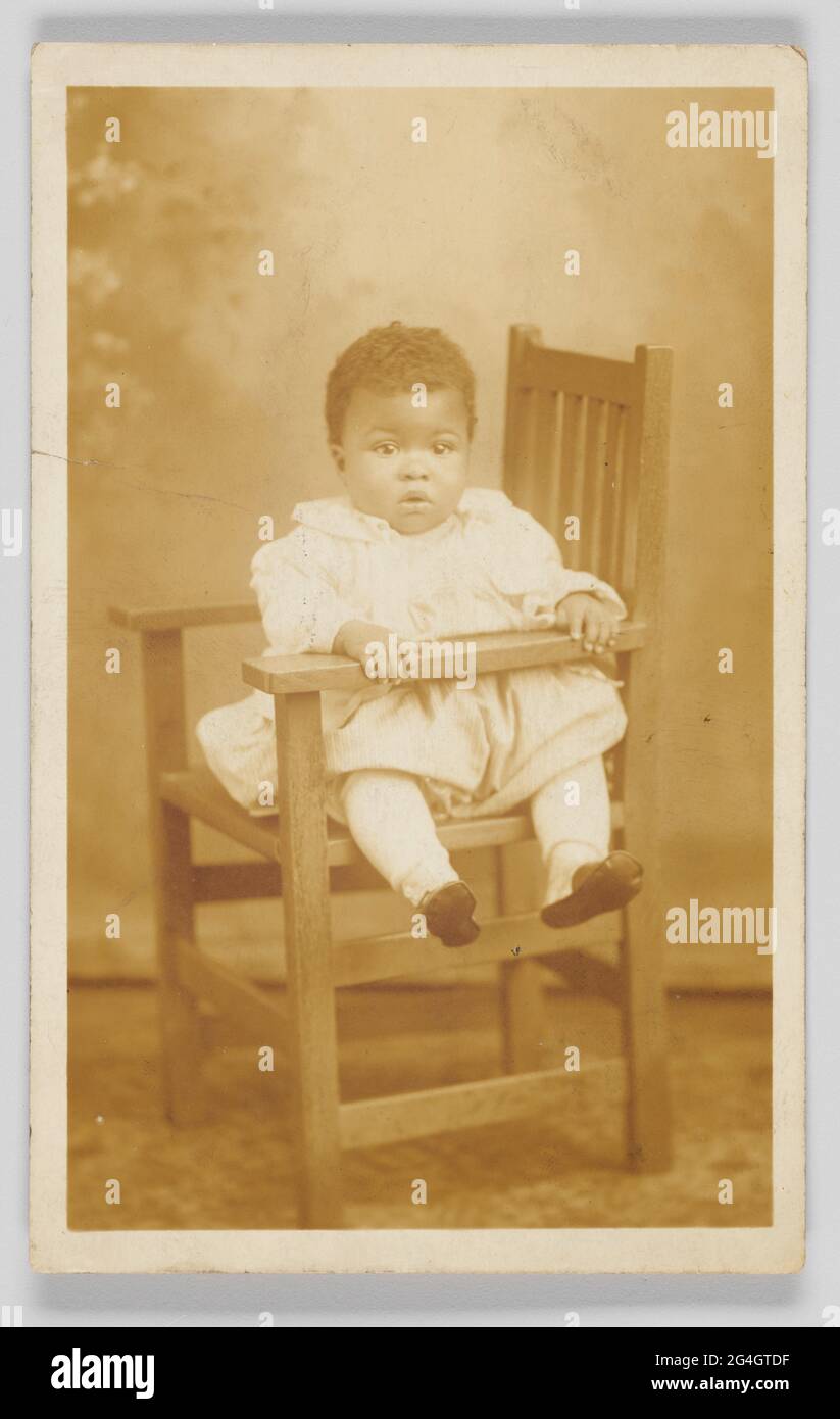 Carte postale photographique d'un enfant non identifié. L'enfant est exposé face à la caméra et assis dans une chaise en bois. La chaise est tournée latéralement, le siège étant orienté vers la gauche de l'image. L'enfant est posé assis vers le bras de la chaise. Les mains de l'enfant reposent sur le bras de la chaise et les jambes de l'enfant sont tirées par l'ouverture entre le bras de la chaise et le siège de la chaise. L'enfant porte une robe de couleur claire, des bas et des chaussures sombres. Le verso de la carte postale photographique n'est pas utilisé et la [CARTE POSTALE] est imprimée en noir en haut et les espaces vides pour [CORRESPONDANCE] et Banque D'Images