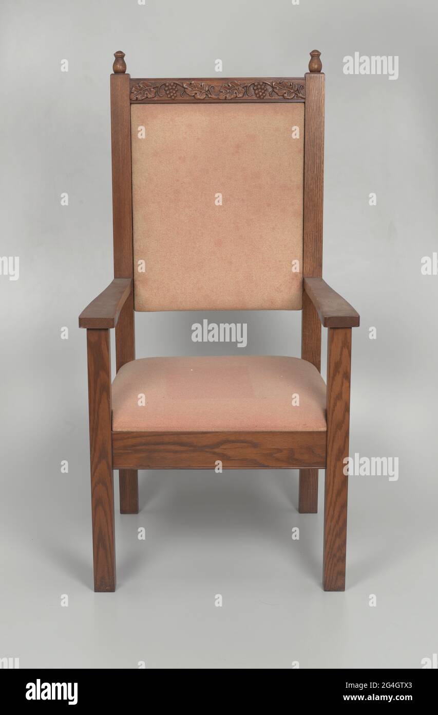 Une chaise à dossier haut avec accoudoirs. La chaise est en bois sombre.  Deux embouts simples se trouvent sur le dessus de la chaise, de chaque  côté. Au sommet de la chaise