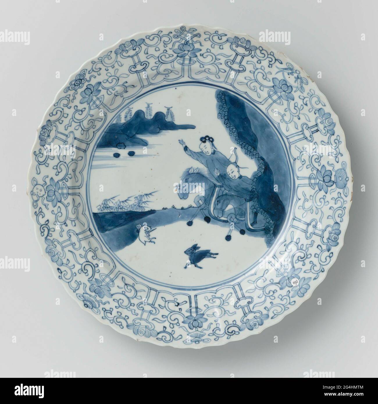 . Plat en porcelaine à paroi ronde et striée et bord festonné, peint en bleu sous-glaçure. Sur l'appartement une scène de yacht avec deux cavaliers armés dans un paysage, jaming sur un lièvre; le bord avec un pneu concerté de lotus boissons, entre les huit symboles chanceux (chakra, coquillages, parasol, verrière, lotus, Vase, poisson, nœud infini); le dos avec seize branches de fleur en vrac. Marqué sur le dessous avec la marque de six caractères de l'empereur Chenghua dans un double cercle. Bord légèrement endommagé. Bleu blanc. Banque D'Images