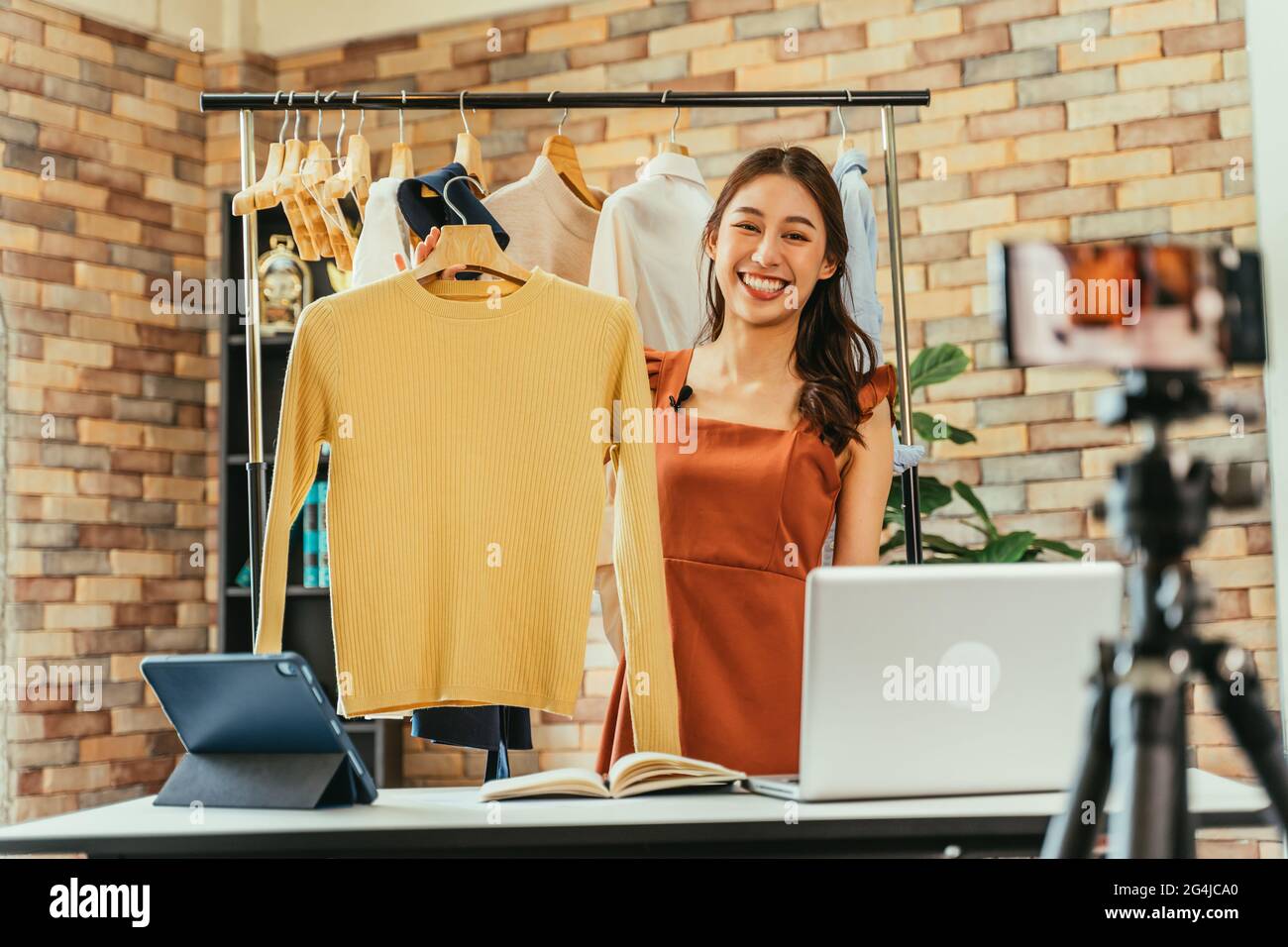 Jeune et belle femme asiatique blogueuse montrant des vêtements devant l'appareil photo de smartphone tout en enregistrant la vidéo vlog et la diffusion en direct dans sa boutique Banque D'Images