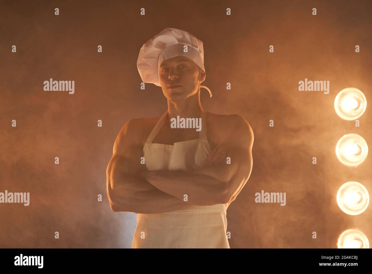 Portrait d'un chef musclé portant un tablier blanc et un chapeau de chef, posant avec des bras pliés sur un fond fumé femme au foyer mâle. Mari dans la cuisine. Brutal mais Banque D'Images