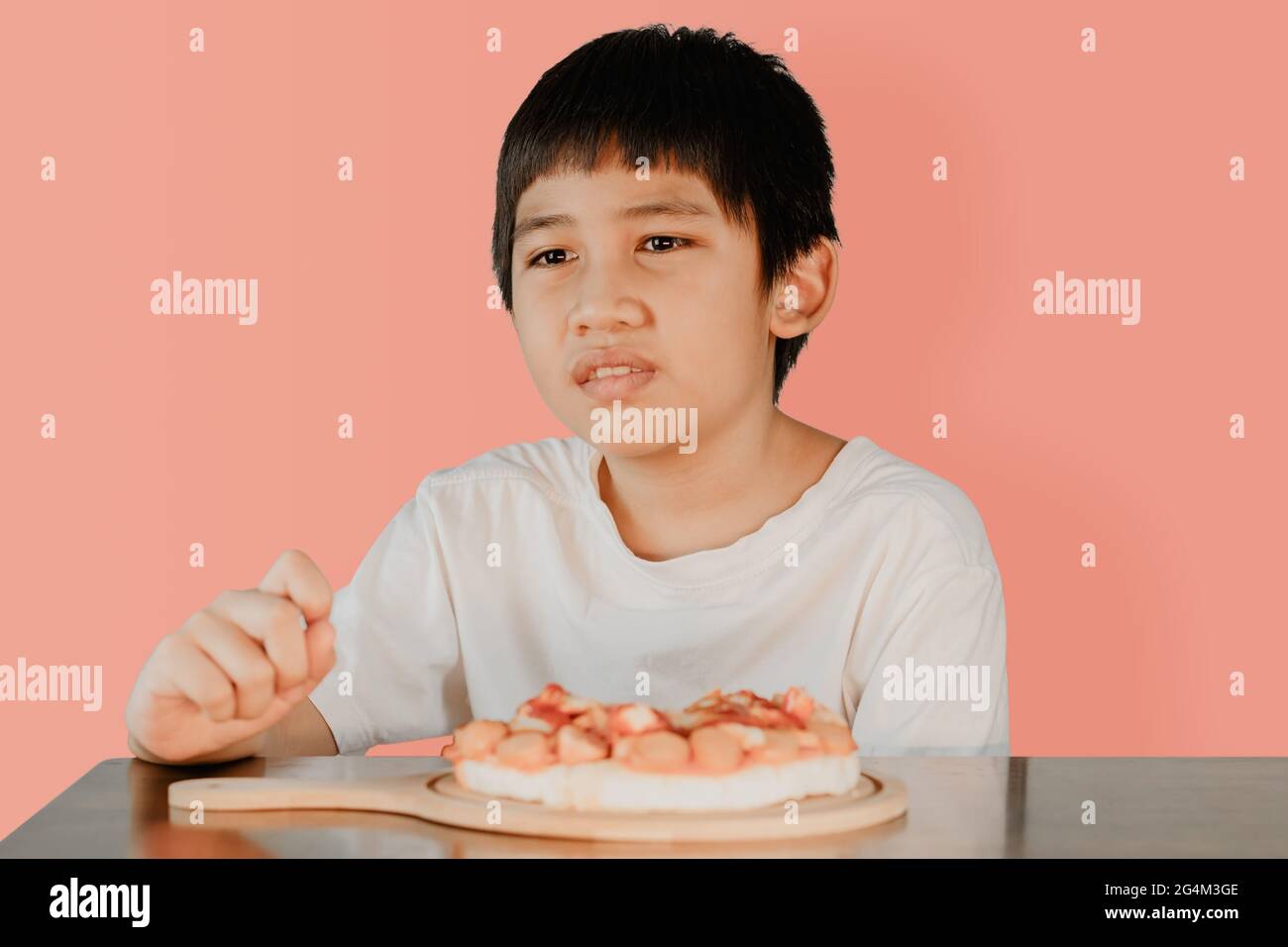 Petit garçon asiatique assis à la table à manger avec une pizza sur la table à l'avant sur fond rose. Banque D'Images Petit garçon asiatique assis à la table à manger avec une pizza sur la table à l'avant sur fond rose. Banque D'Images