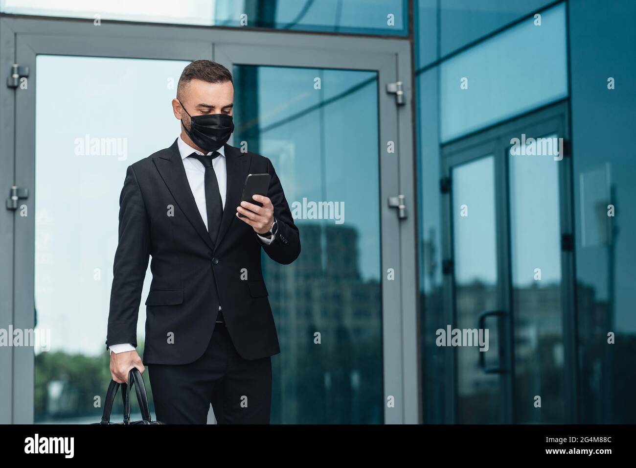 Entrepreneur mature dans un masque facial utilisant un smartphone se tenant près du centre de bureau dans la ville urbaine, espace libre Banque D'Images