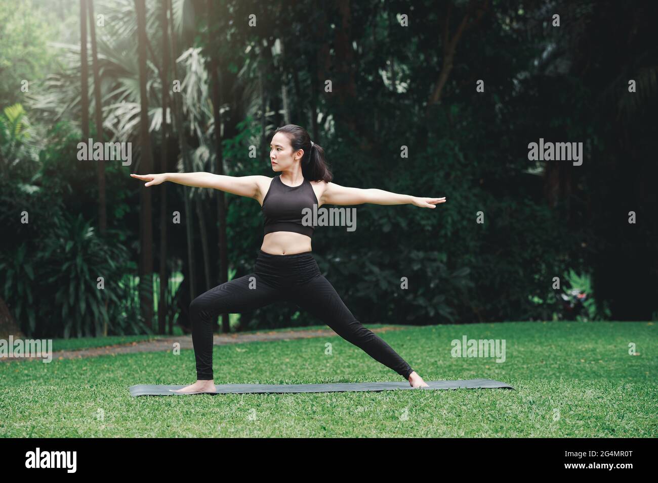 Femme asiatique pratiquant le yoga en posture du guerrier (Virabhadrasana) sur le tapis dans le parc extérieur. Banque D'Images