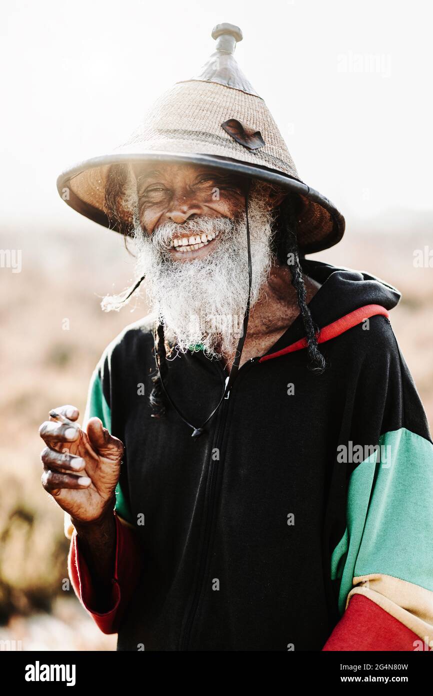 rastafari ethnique ancien gai avec des dreadlocks regardant l'appareil-photo tout en fumant de l'herbe debout dans un pré sec dans la nature Banque D'Images