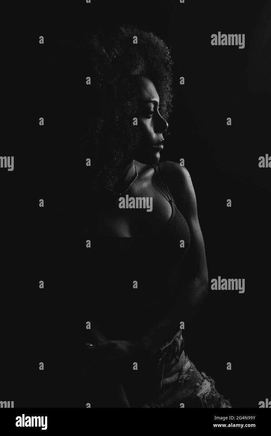 Une femme afro-américaine aux cheveux bouclés, avec un petit top tendance et un Jean à la main sur la taille, sur fond noir, en studio Banque D'Images Une femme afro-américaine aux cheveux bouclés, avec un petit top tendance et un Jean à la main sur la taille, sur fond noir, en studio Banque D'Images