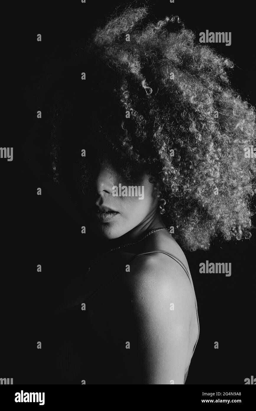 Noir et blanc charmant modèle africain américain avec cheveux bouclés regardant l'appareil photo dans le studio sombre Banque D'Images Noir et blanc charmant modèle africain américain avec cheveux bouclés regardant l'appareil photo dans le studio sombre Banque D'Images