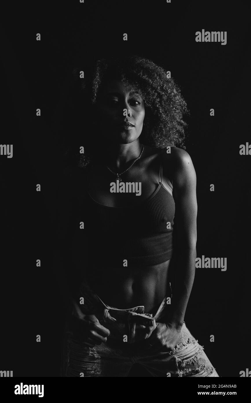 Une femme afro-américaine aux cheveux bouclés, avec un petit top tendance et un Jean à la main sur la taille, sur fond noir, en studio Banque D'Images Une femme afro-américaine aux cheveux bouclés, avec un petit top tendance et un Jean à la main sur la taille, sur fond noir, en studio Banque D'Images