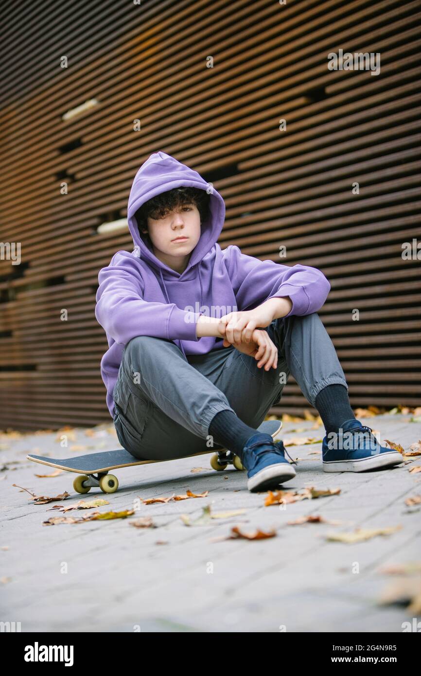 Tout le corps d un adolescent dans des vetements decontractes avec un sweat a capuche regardant l appareil photo tout en etant assis sur un skateboard au mur dans la rue avec des feuilles