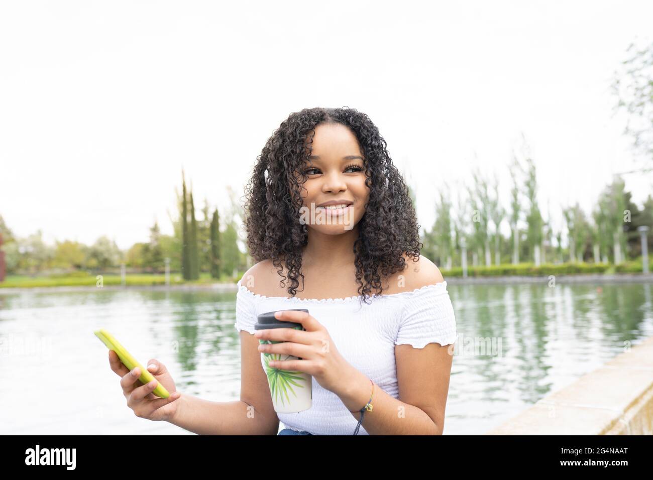 Jeune femme afro-américaine positive en vêtements d'été avec tasse de café à emporter et téléphone près de la rivière et des arbres verts dans le parc pendant la journée tandis que looki Banque D'Images