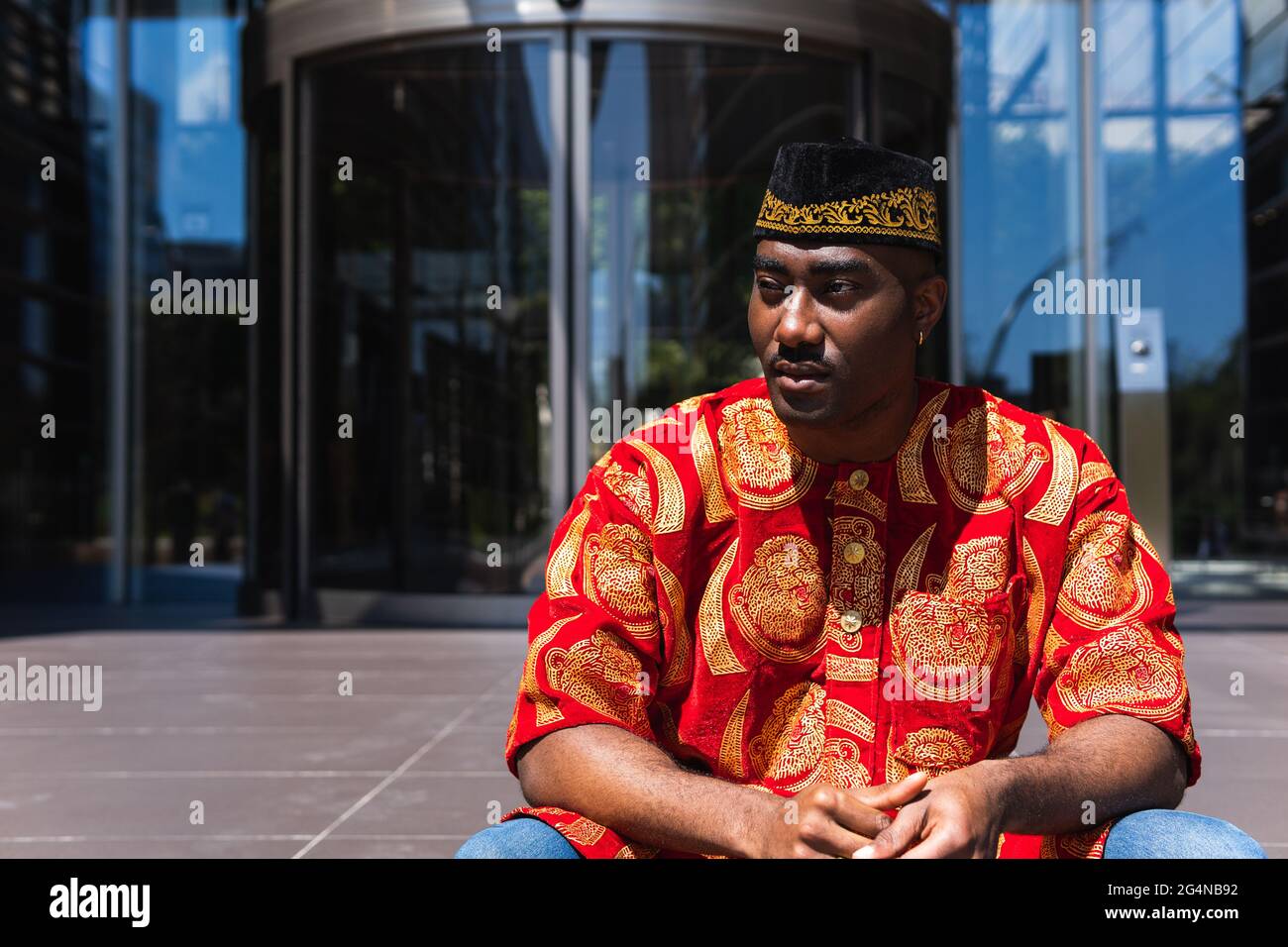 Un homme africain adulte sérieux en vêtements rouges traditionnels et  casquette kufi qui regarde loin tout en étant assis dans la rue de la ville  près du bâtiment avec des murs de