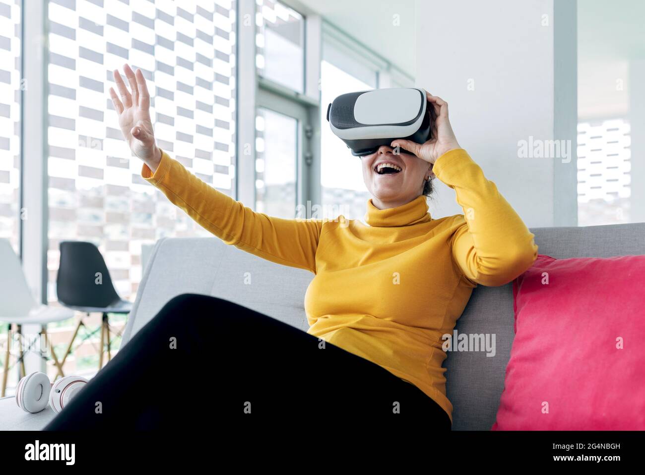 Femme dans une tenue décontractée assise sur un canapé avec oreillers tout en utilisant des lunettes VR près des écouteurs et des fenêtres dans un bâtiment lumineux Banque D'Images