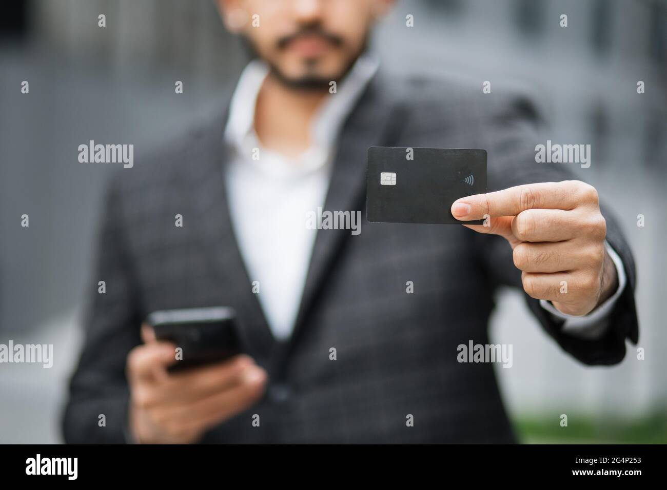 Gros plan de l'homme musulman dans des vêtements formels debout dans la rue de la ville et montrant la carte de crédit noire. Homme occupé utilisant un téléphone cellulaire moderne pour faire des achats en ligne à l'extérieur. Banque D'Images