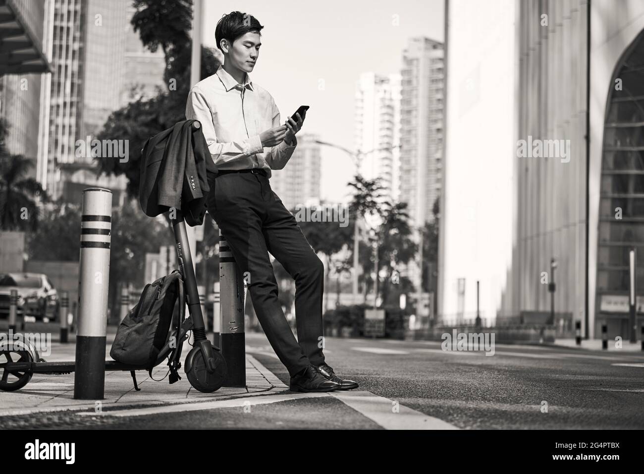 jeune homme d'affaires asiatique debout à côté de son scooter électrique dans la rue dans le détracteur de la ville moderne, noir et blanc Banque D'Images