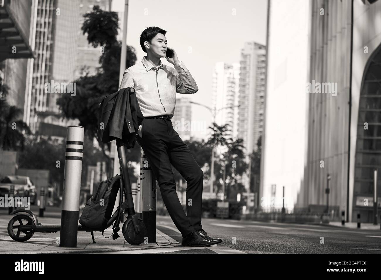 un jeune homme d'affaires asiatique se tenant à côté de son scooter électrique dans la rue en faisant un appel avec un téléphone portable dans le détracteur de la ville moderne, noir et blanc Banque D'Images