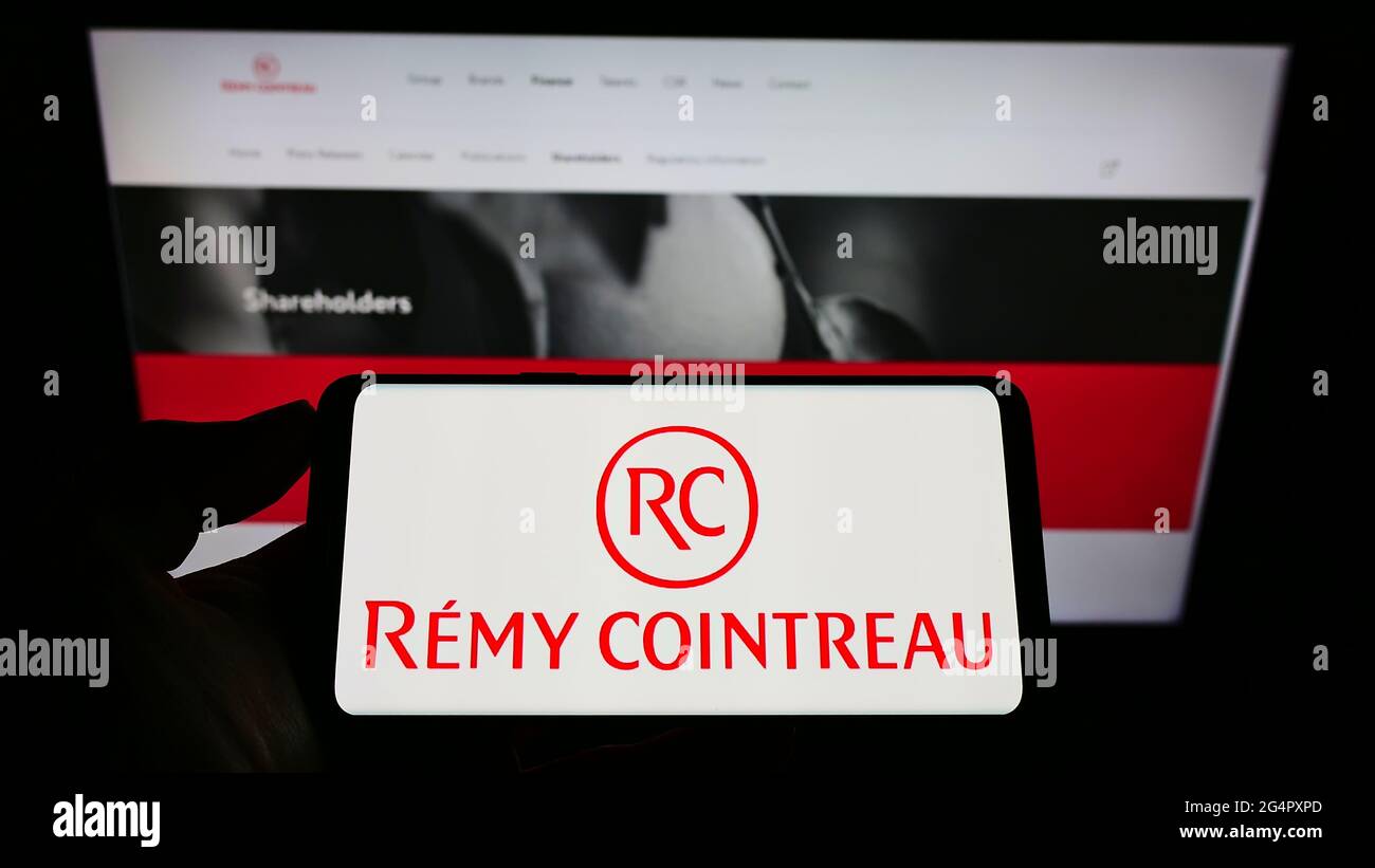Personne tenant un téléphone portable avec le logo de la société française de spiritueux Remy Cointreau S.A. sur écran en face de la page web d'affaires. Mise au point sur l'affichage du téléphone. Banque D'Images