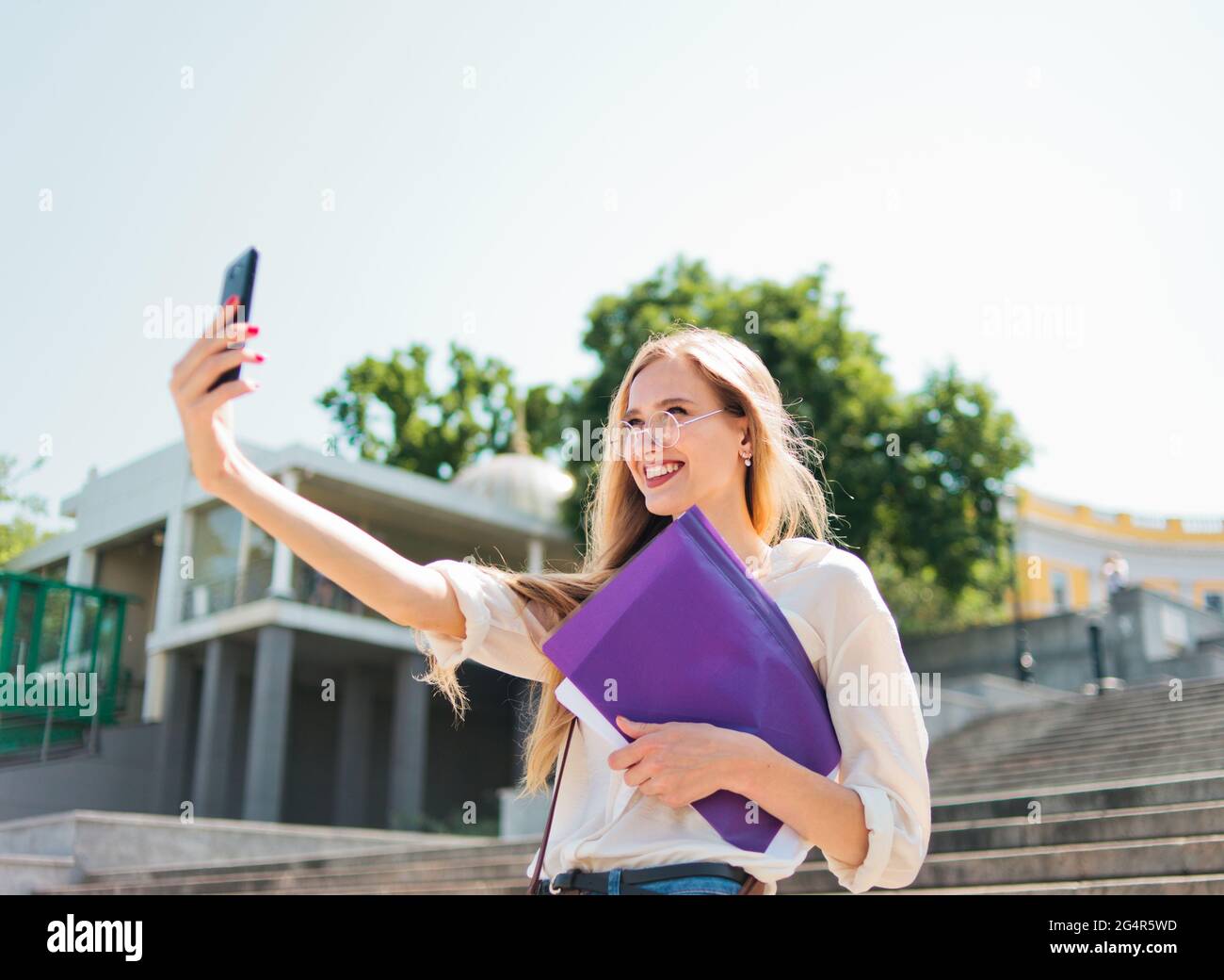 Jeune femme caucasienne étudiant dans des vêtements décontractés avec l'esse dans dossier faisant selfie sur le téléphone en descendant les escaliers Banque D'Images
