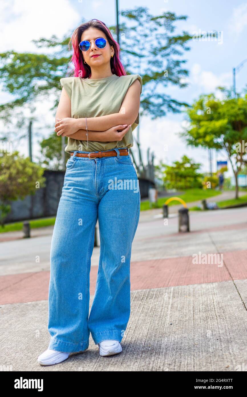 photographie verticale d'une femme debout avec ses bras croisés, portant des lunettes de soleil bleues et regardant l'appareil photo Banque D'Images