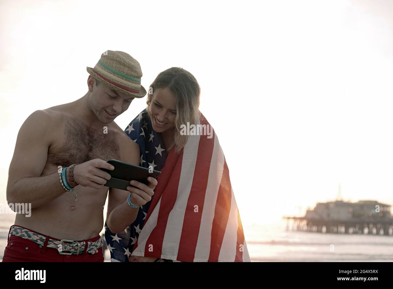 Beau homme utilisant la tablette numérique par petite amie enveloppé dans le drapeau sur la plage de Santa Monica Banque D'Images