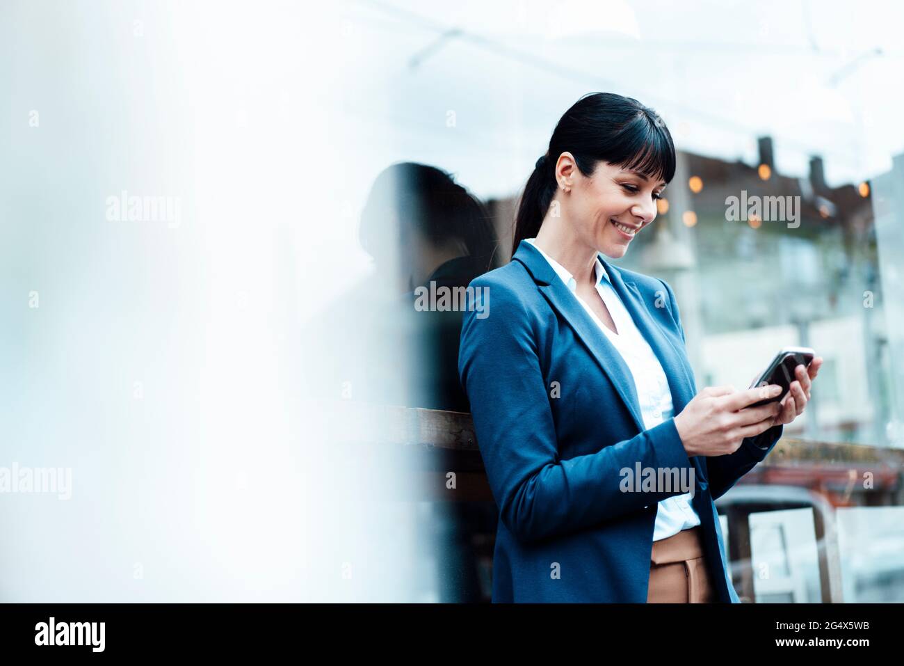 Femme d'affaires souriante utilisant un téléphone portable tout en se penchant sur la fenêtre du café Banque D'Images