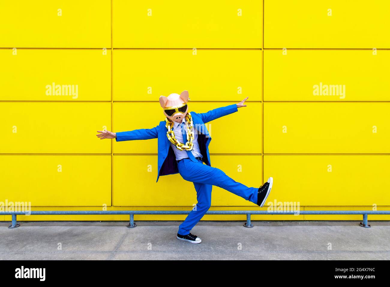 Homme portant un costume bleu vif, un masque de porc et une grande chaîne dorée debout sur une jambe devant le mur jaune Banque D'Images