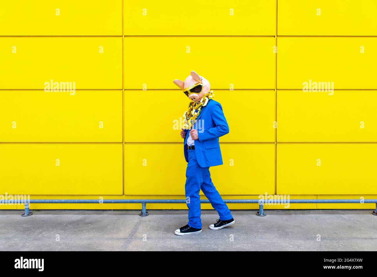 Homme portant un costume bleu vif, un masque de porc et une grande chaîne dorée posant contre le mur jaune Banque D'Images