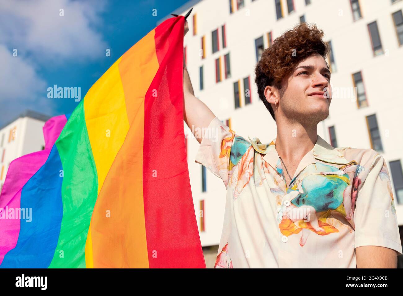 Jeune homme regardant loin tout en soufflant le drapeau arc-en-ciel Banque D'Images