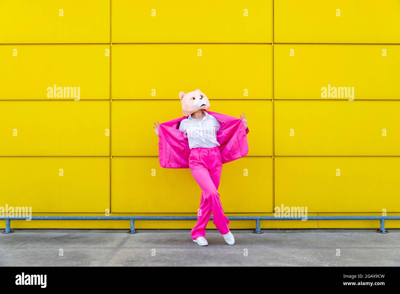 Femme portant un costume rose vif et un masque d'ours debout devant un mur jaune avec une veste de spread Banque D'Images