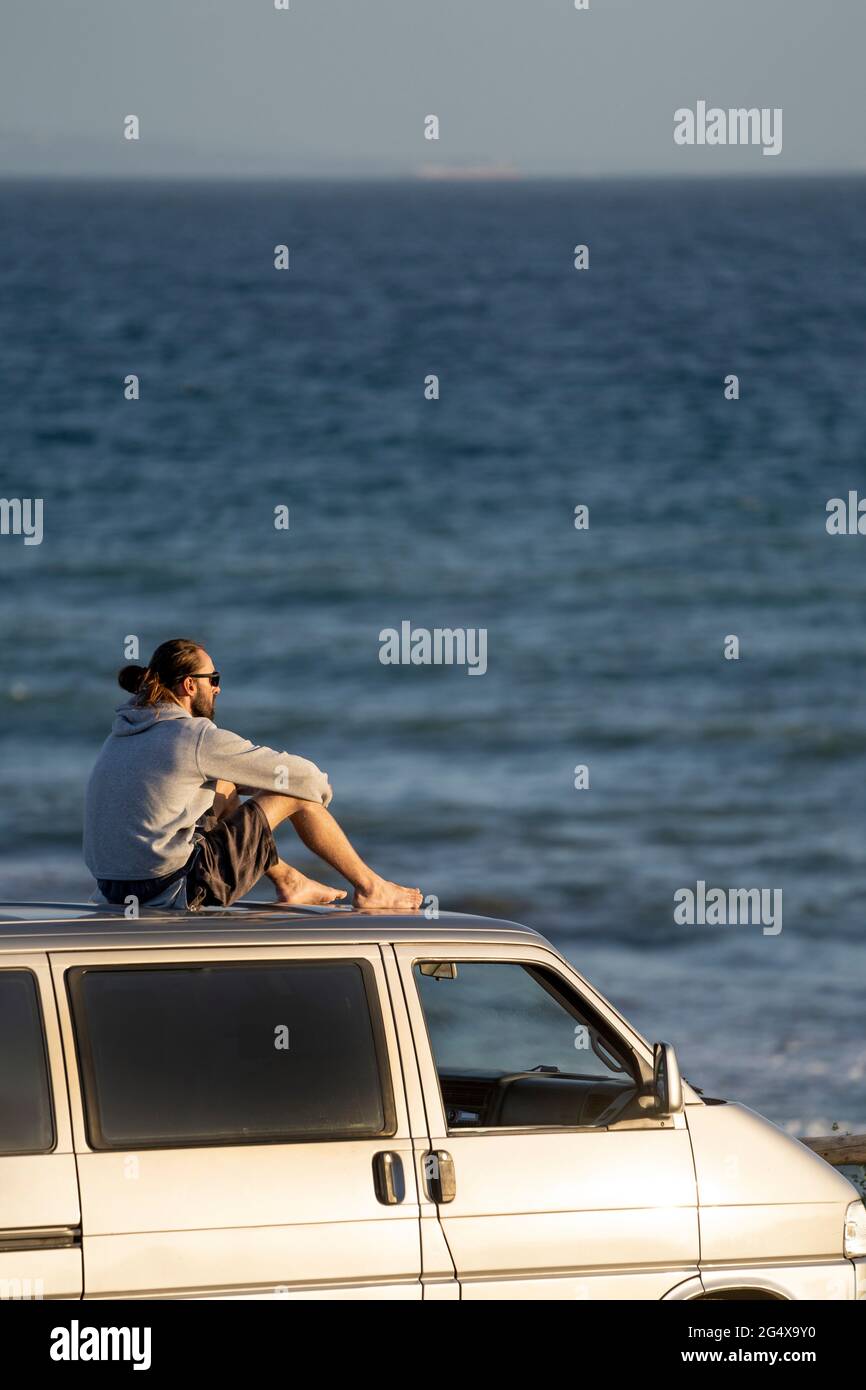 Homme regardant la vue sur la mer tout en étant assis sur le toit de la camionnette Banque D'Images