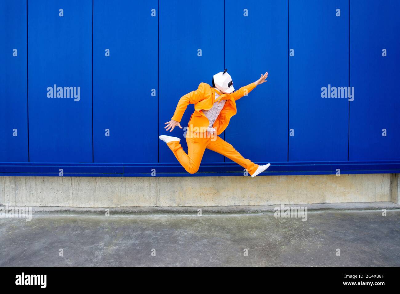 Homme portant un costume orange vif et un masque de panda sautant contre le mur bleu Banque D'Images