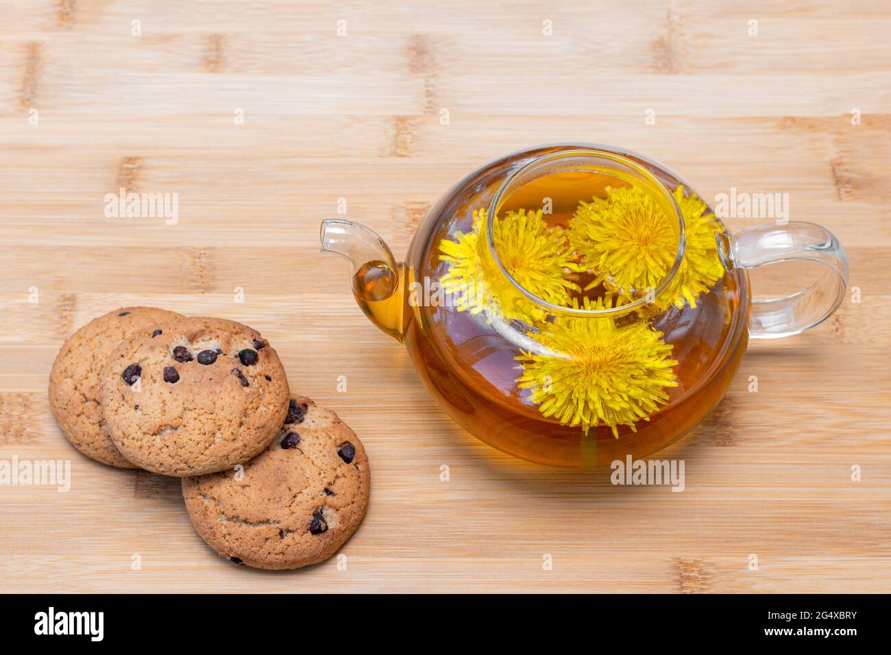 Thé de pissenlit à base de plantes saines dans une théière en verre et des biscuits aux raisins secs sur un arrière-plan en bois. Petit déjeuner d'été. Concept de saine alimentation Banque D'Images