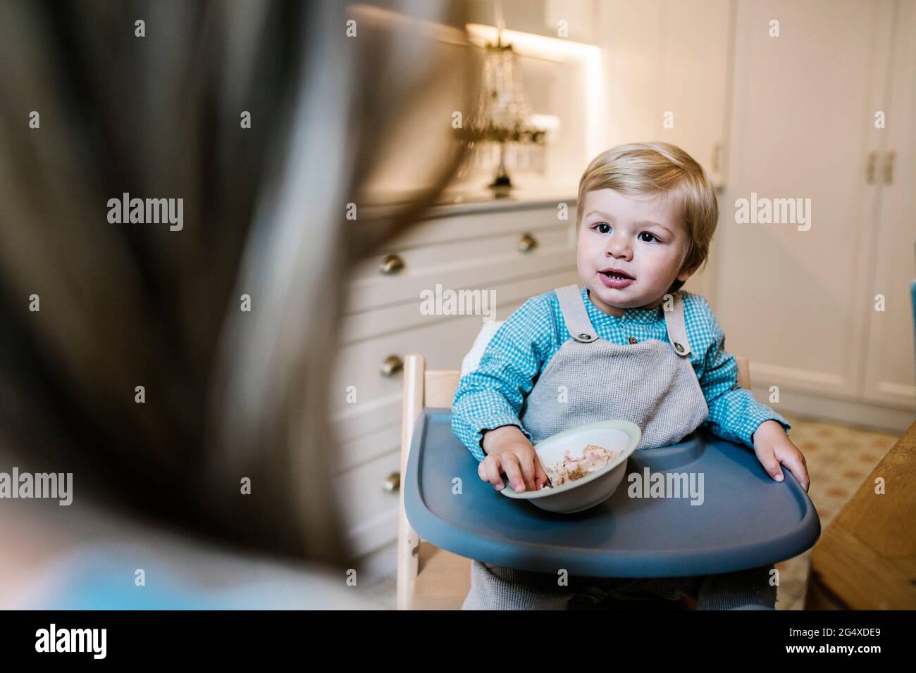 Fils regardant la mère tout en ayant la nourriture sur une chaise haute à la maison Banque D'Images