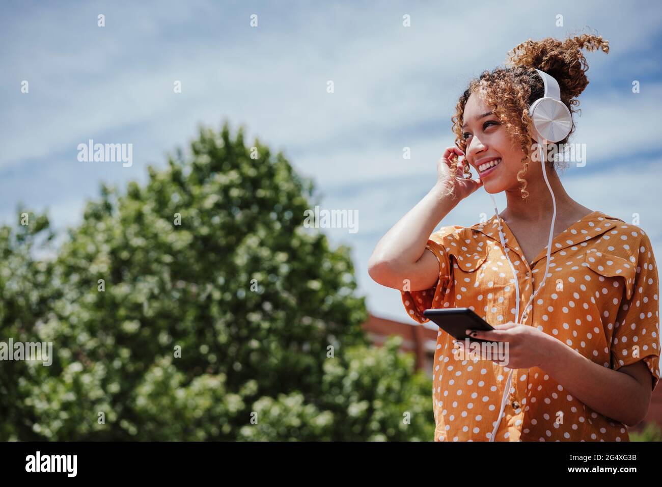 Femme souriante aux cheveux bouclés qui regarde loin tout en écoutant de la musique à travers des écouteurs sur le balcon Banque D'Images