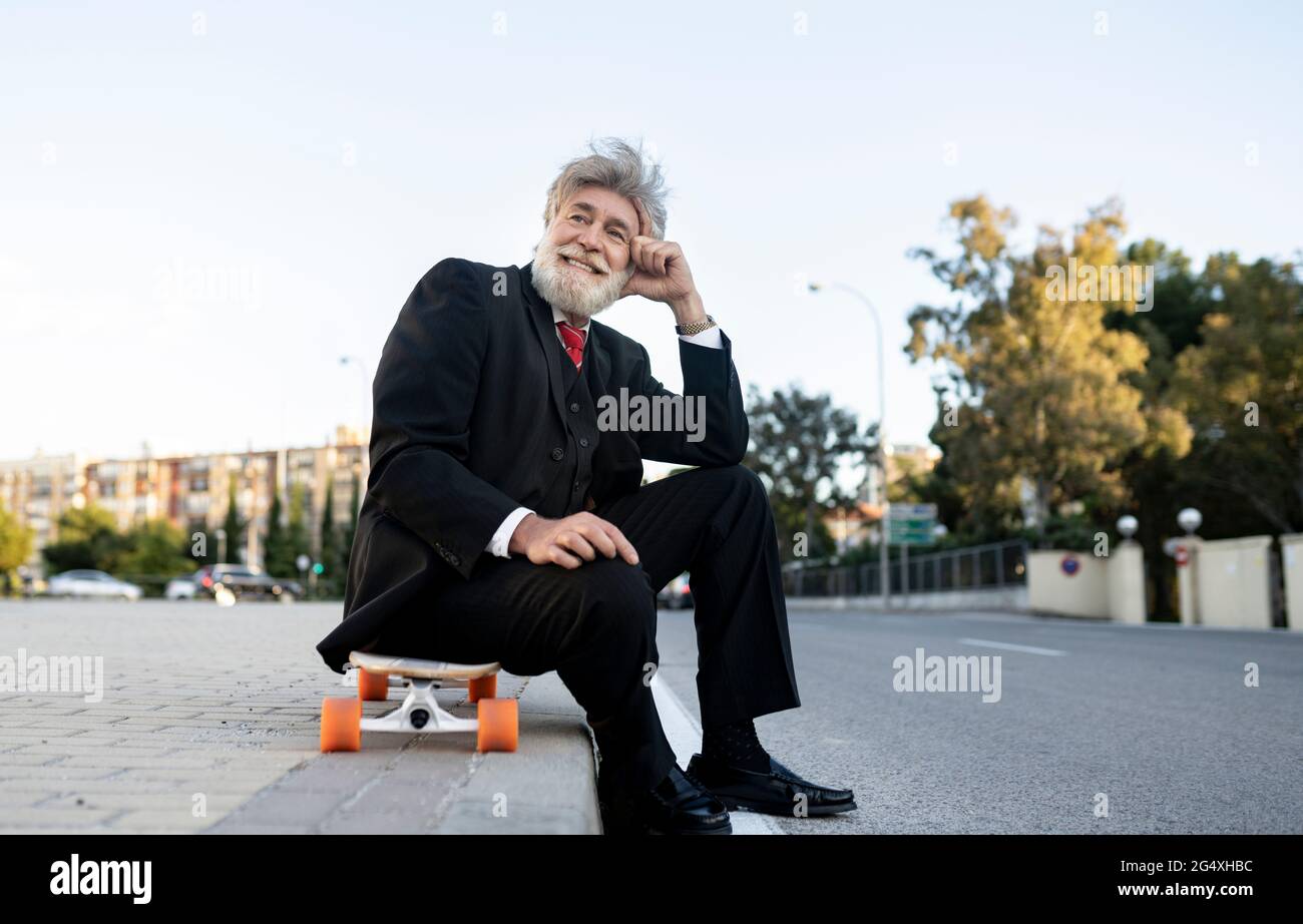 Homme d'affaires souriant et mûr assis sur un skateboard tout en rêvant Banque D'Images