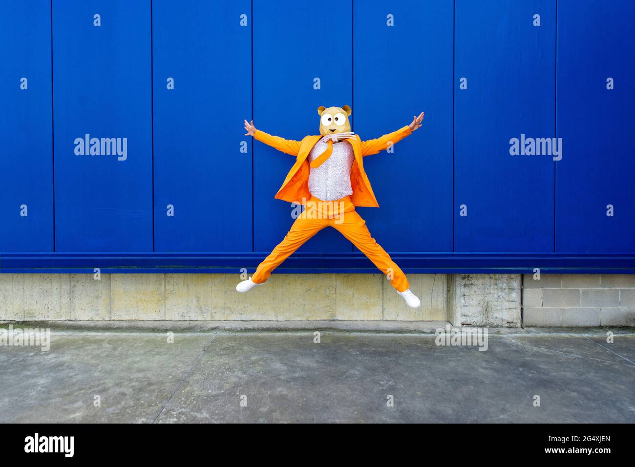 Homme portant un costume orange vif et un masque de rongeur qui saute contre le mur bleu Banque D'Images