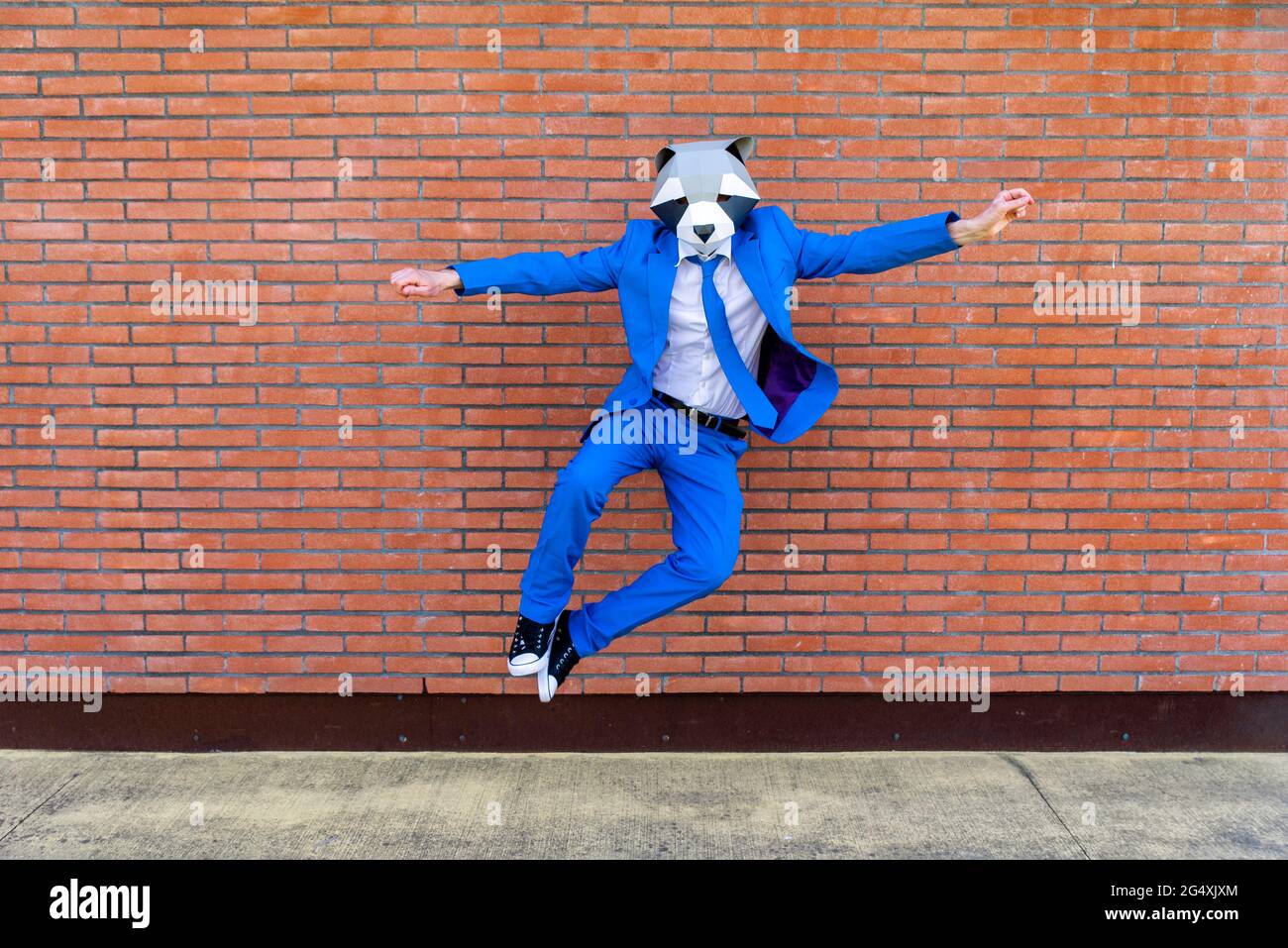 Homme portant un costume bleu vif et un masque de raton laveur qui saute contre un mur de briques Banque D'Images