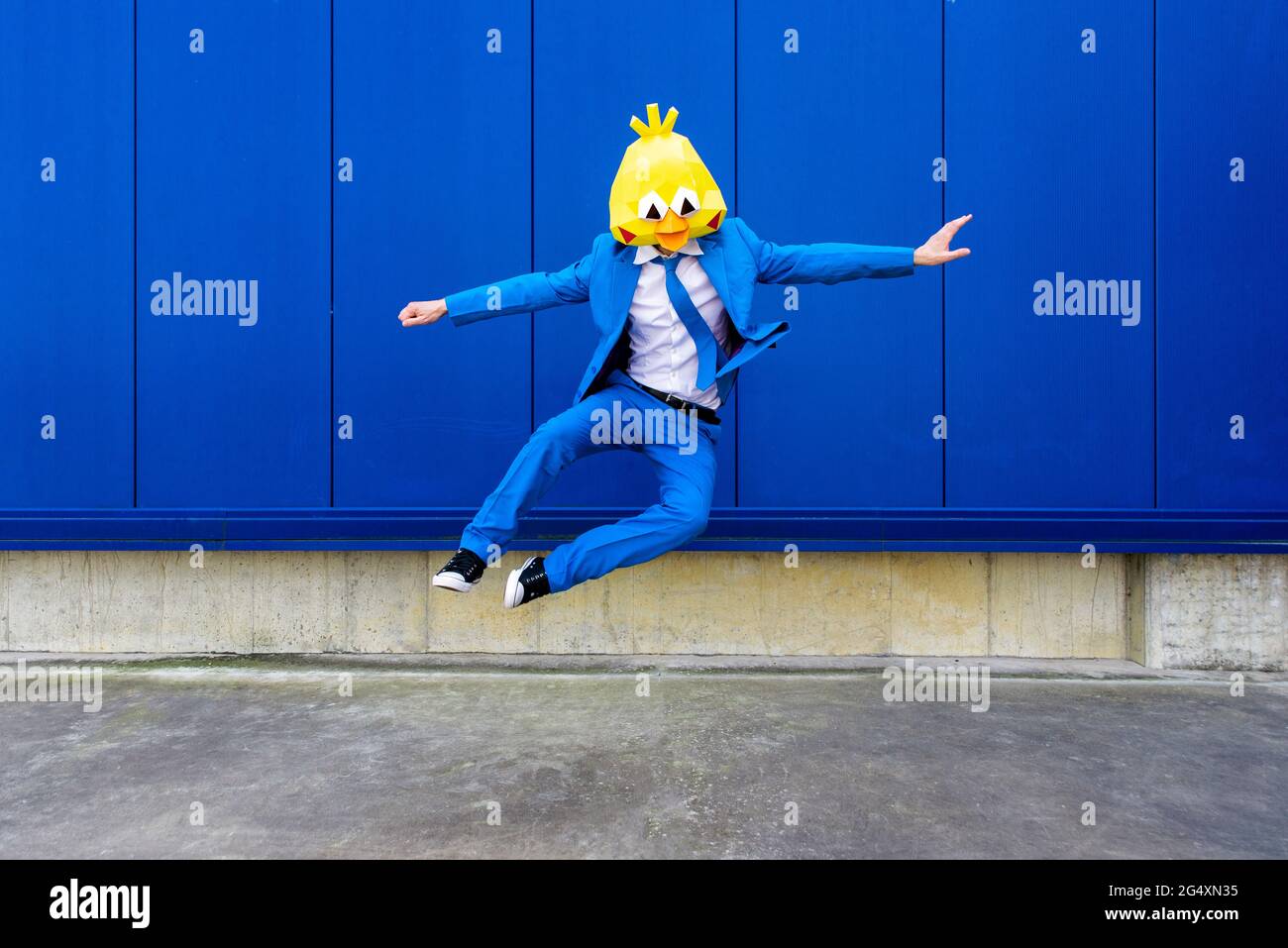 Homme portant un costume bleu vif et un masque d'oiseau sautant contre le mur bleu Banque D'Images