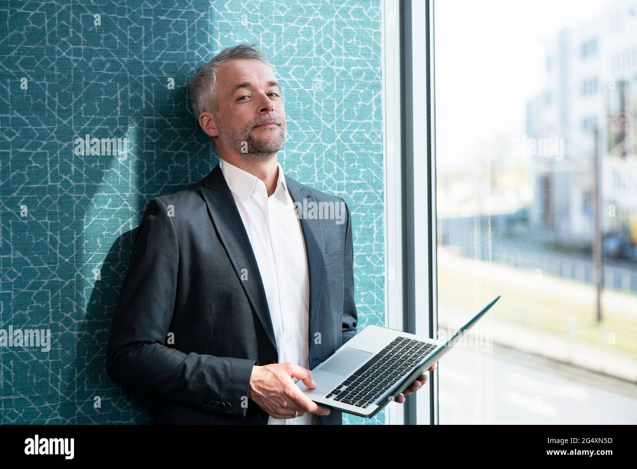 Homme d'affaires mature avec ordinateur portable penché sur le mur près de la fenêtre au bureau Banque D'Images