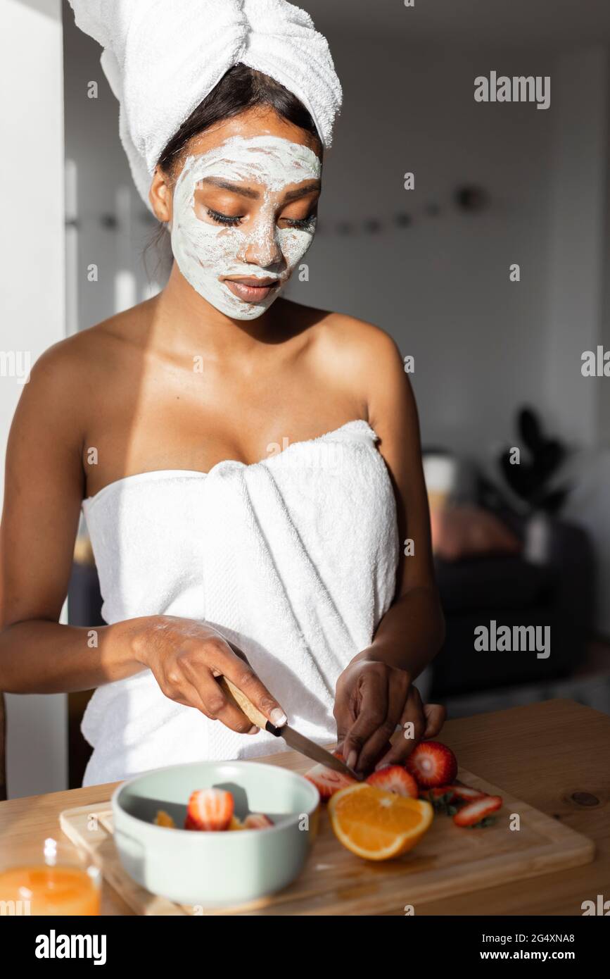 Femme avec masque facial portant une serviette tout en coupant des fruits dans la cuisine à la maison Banque D'Images