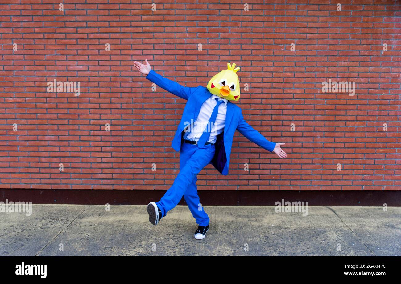 Homme portant un costume bleu vif et un masque d'oiseau posant sur une jambe devant le mur de briques Banque D'Images