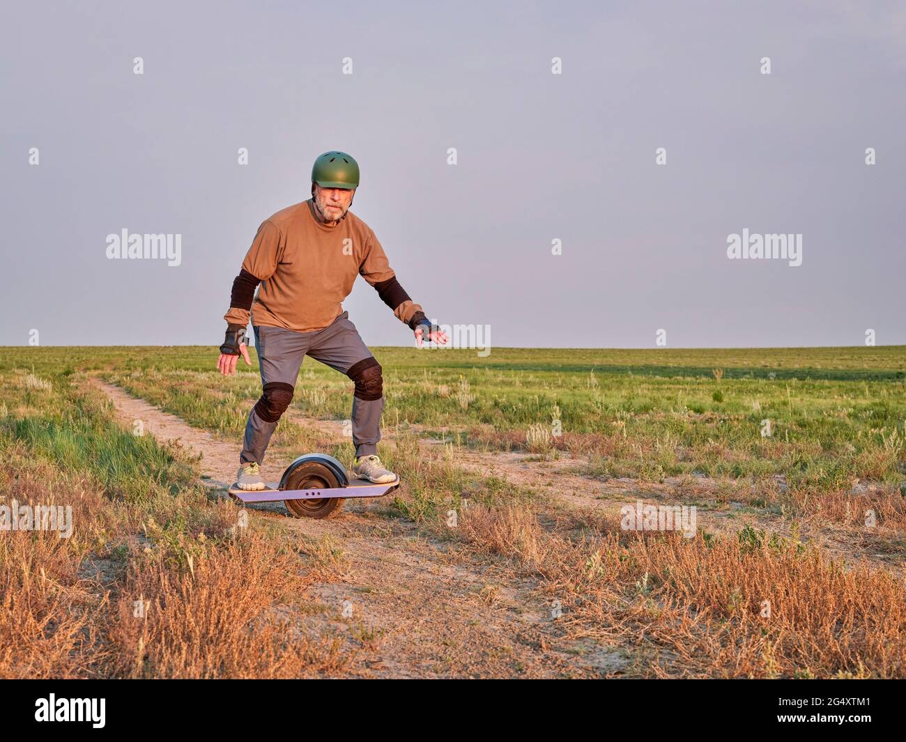 promenade au lever du soleil sur une prairie, un homme âgé d'âge mûr est à cheval sur un skateboard électrique à une roue sur une route de terre Banque D'Images